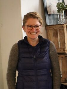woman with blonde hair tied wearing glasses grey sweater and blue vest smiling to the right of the camera